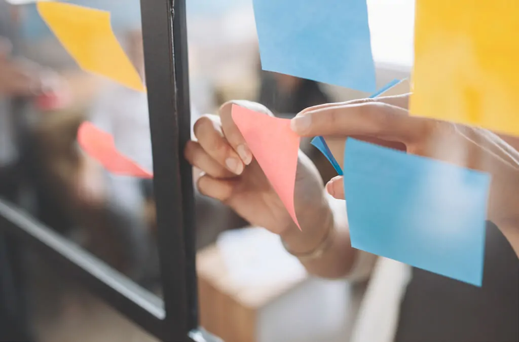 Close-up of a hand placing colorful sticky notes on a glass board during a brainstorming session.