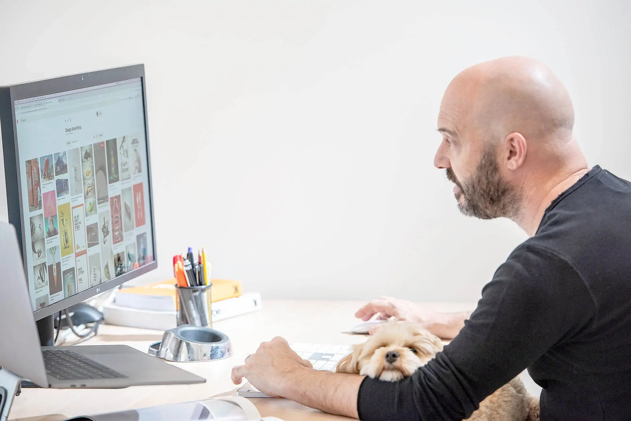 Man working at a desk with a computer, accompanied by a small dog resting on his arm.