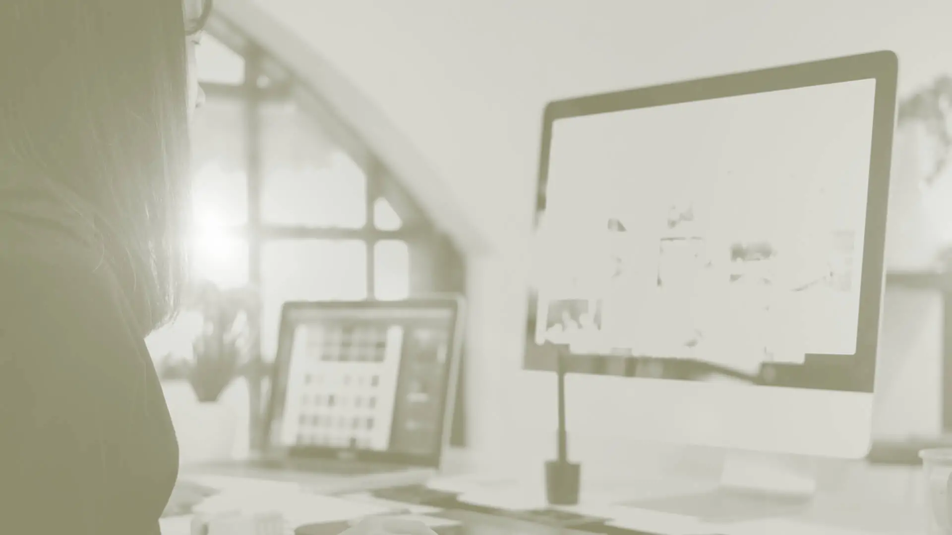 Person working at a desk with a computer and laptop in a softly lit workspace.